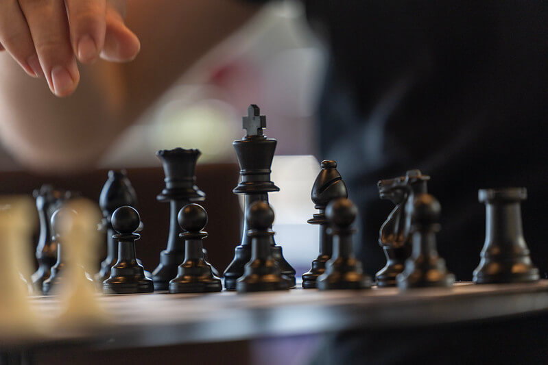 A hand reaches to move a black chess piece on a wooden board, with other pieces visible in the foreground.
